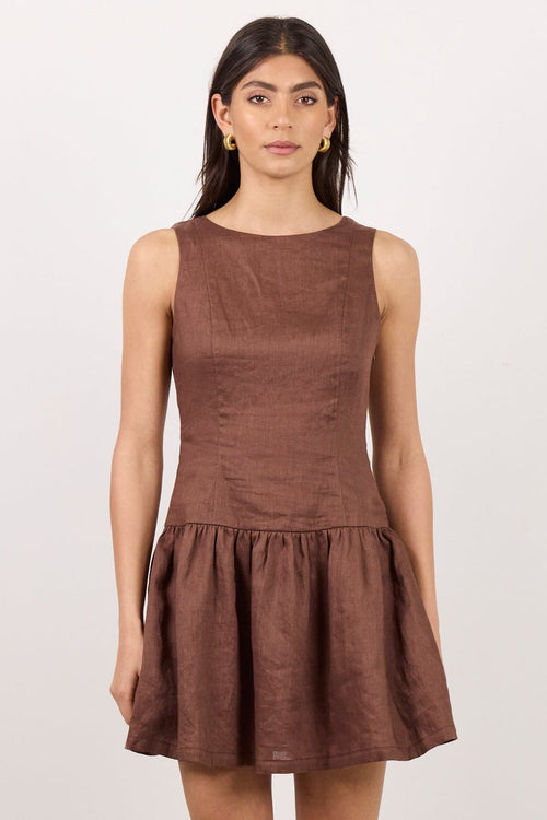 A woman modeling a sleeveless brown mini dress with a gathered peplum hem, paired with black sandals, standing against a plain white studio background.