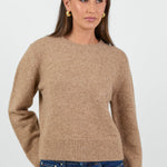 A woman wearing a soft, biscuit-colored crew neck jumper with long sleeves and ribbed cuffs, paired with dark blue high-waisted jeans.