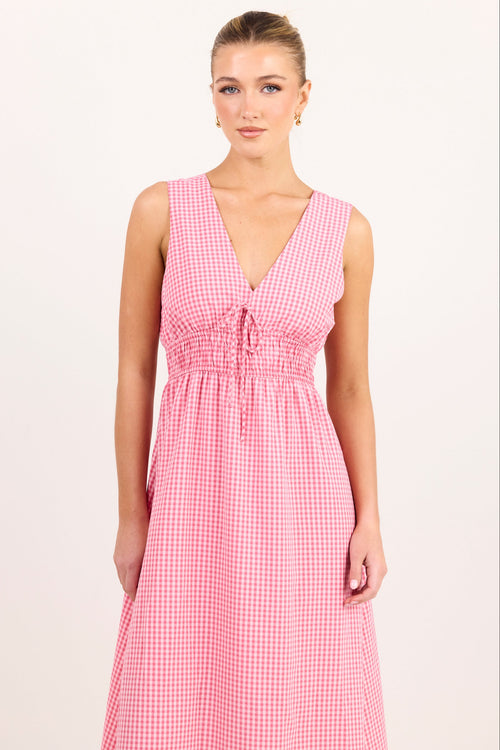 A woman modeling a sleeveless pink and white gingham maxi dress with a smocked waist and tie-front neckline, photographed against a plain light background.