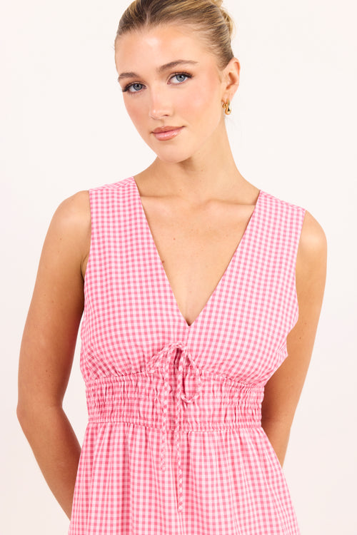 A woman wearing a pink and white gingham sleeveless dress with a deep V-neckline and a smocked waist with a front tie, set against a plain light background.