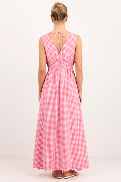 A woman viewed from the back wearing a sleeveless pink gingham maxi dress with a tie-strap open back, standing against a plain white studio background.