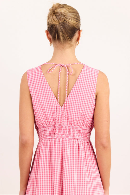 A woman viewed from the back wearing a sleeveless pink and white gingham dress with a tie-back neckline and a smocked waist, against a plain light background.