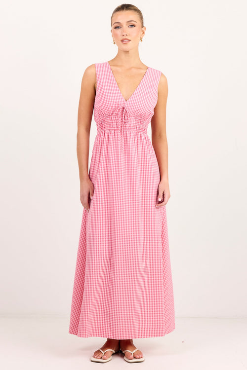 A woman modeling a sleeveless pink and white gingham maxi dress with a V-neckline and gathered waist, standing against a plain white studio background.