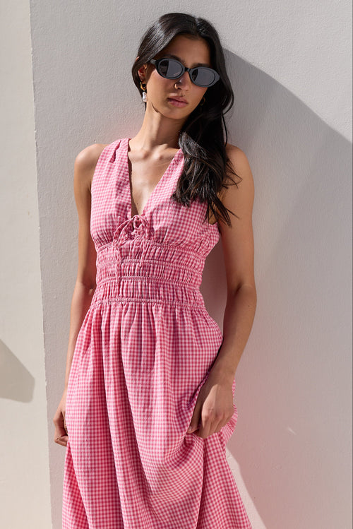 A woman wearing a pink and white gingham smocked dress and dark sunglasses, posing against a white stucco wall.