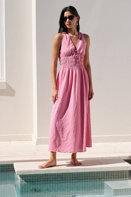 A woman wearing a sleeveless pink maxi dress and sunglasses stands barefoot on the edge of a swimming pool next to a white stucco building.