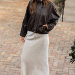 A woman stands on a cobblestone courtyard wearing a black leather bomber jacket, a long cream-colored midi skirt, and black flip-flop sandals.