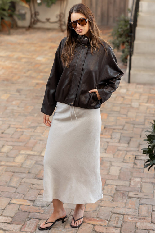 A woman stands on a cobblestone courtyard wearing a black leather bomber jacket, a long cream-colored midi skirt, and black flip-flop sandals.