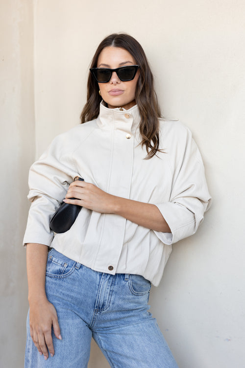 A woman wearing a cream-colored high-neck jacket, blue jeans, and black sunglasses, holding a black clutch bag against a neutral beige wall.