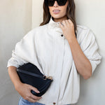 A woman wearing a cream-colored high-neck button-up jacket, blue jeans, and black sunglasses, holding a black clutch bag against a neutral beige wall.