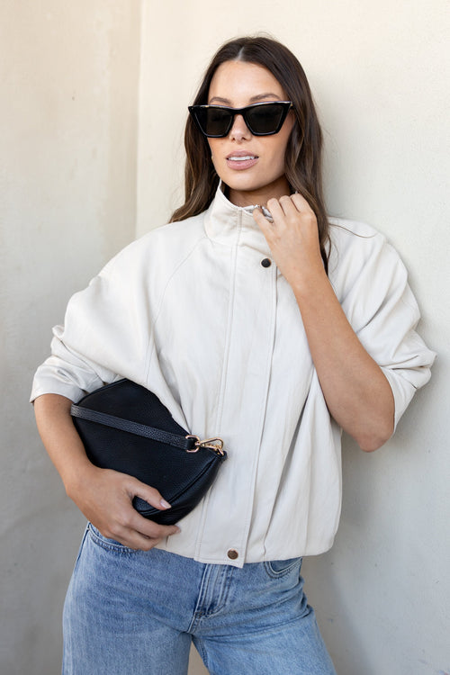 A woman wearing a cream-colored high-neck button-up jacket, blue jeans, and black sunglasses, holding a black clutch bag against a neutral beige wall.