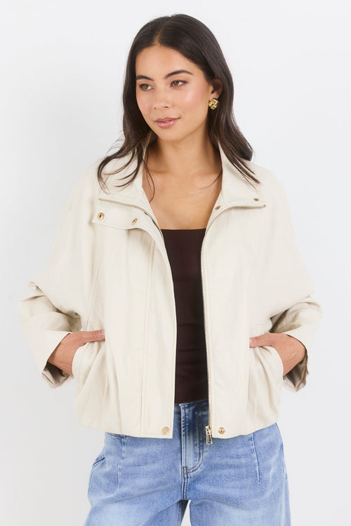 Woman wearing a cream jacket, black top, and blue jeans on a white background