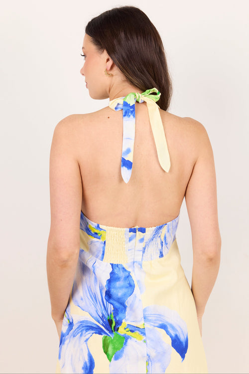 A woman shown from the back wearing a halter dress with a pale yellow base and a blue floral print. The dress features a tie-neck detail with blue and green watercolor-style accents.