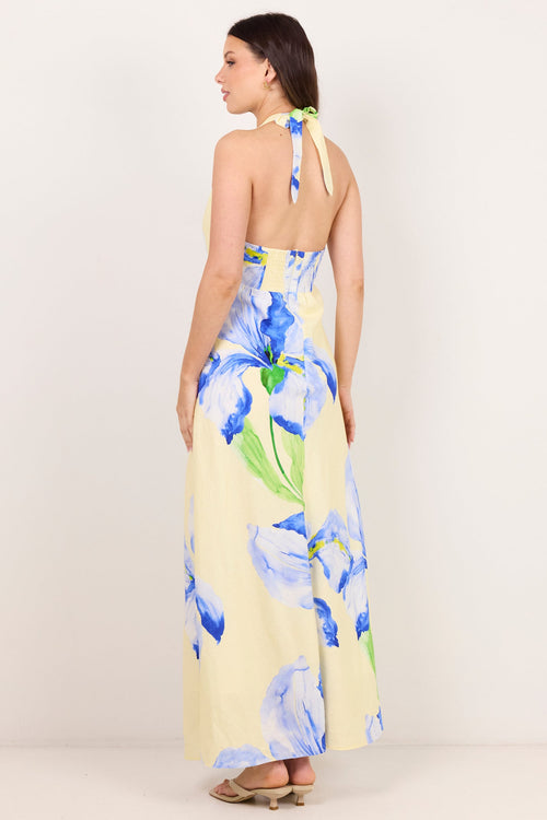 A woman modeling a halter-neck maxi dress with a blue floral print on a pale yellow background, shown from the back against a plain off-white studio backdrop.