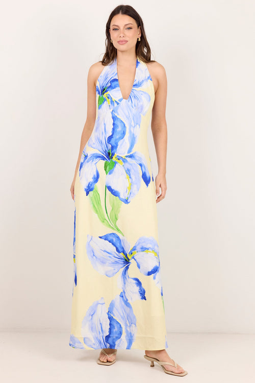 A woman modeling a sleeveless halter-neck maxi dress with a large blue iris floral print on a pale yellow background, standing against a plain off-white studio backdrop.