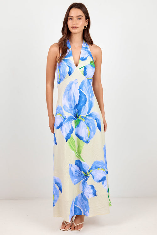 A woman modeling a sleeveless halter-neck maxi dress with a large blue floral print on a cream background, standing against a plain white studio backdrop.