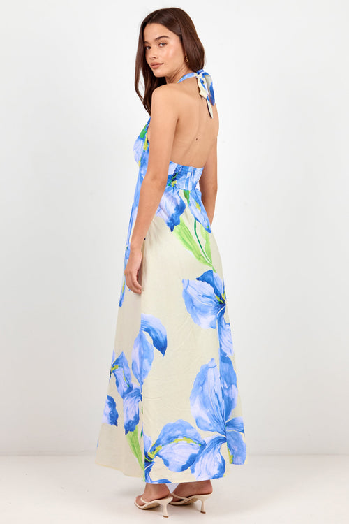 A woman modeling a halter-neck maxi dress with a large blue floral print on a cream background, shown from the back against a plain white studio backdrop.