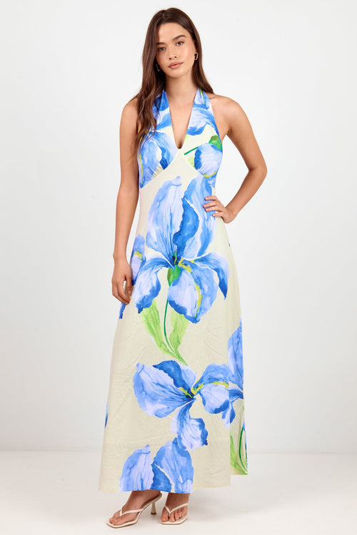 A woman modeling a sleeveless halter-neck maxi dress with a large blue floral print on a cream background, standing against a plain light gray studio backdrop.