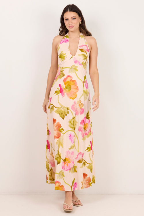 A woman modeling a sleeveless halter-neck maxi dress with a vibrant pink, orange, and green floral print on a cream background, set against a plain off-white studio backdrop.