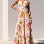 A woman models a sleeveless halter-neck maxi dress with a pink, green, and yellow floral watercolor print, standing on the edge of a swimming pool against a white stucco wall.