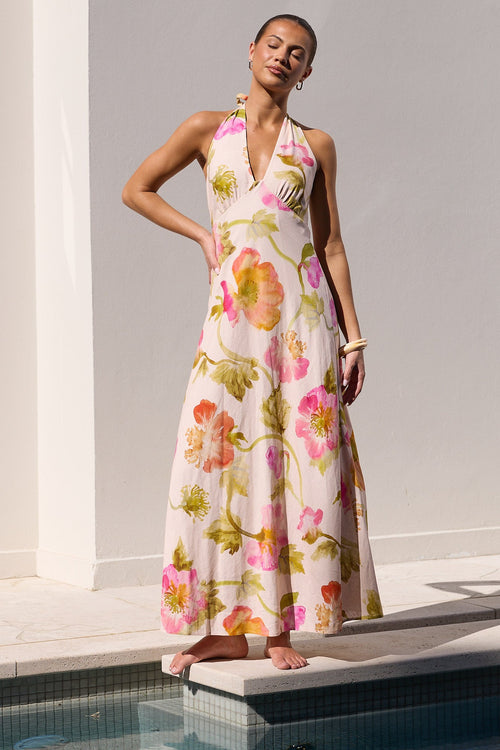A woman models a sleeveless halter-neck maxi dress with a pink, green, and yellow floral watercolor print, standing on the edge of a swimming pool against a white stucco wall.