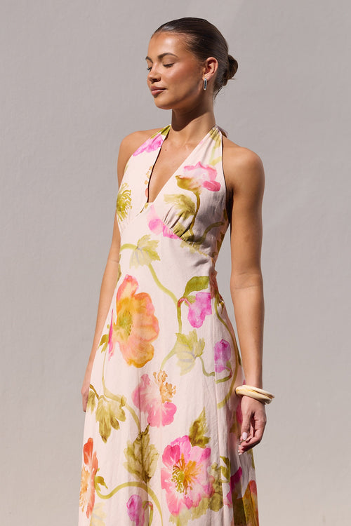 A woman wearing a halter-neck maxi dress with a vibrant pink, orange, and green floral print on a light beige background, standing against a plain light gray wall.