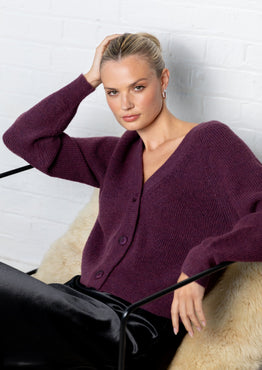 Woman wearing a purple cardigan and black pants sitting on a chair with a white brick wall background