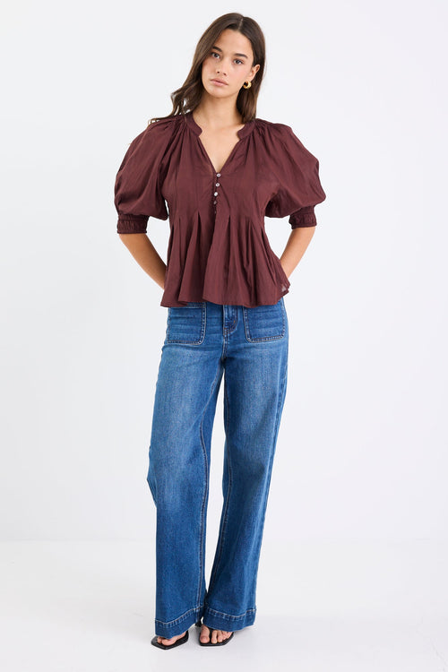 Woman wearing a chocolate brown pleated short-sleeve top with a button-down front, paired with blue jeans.
