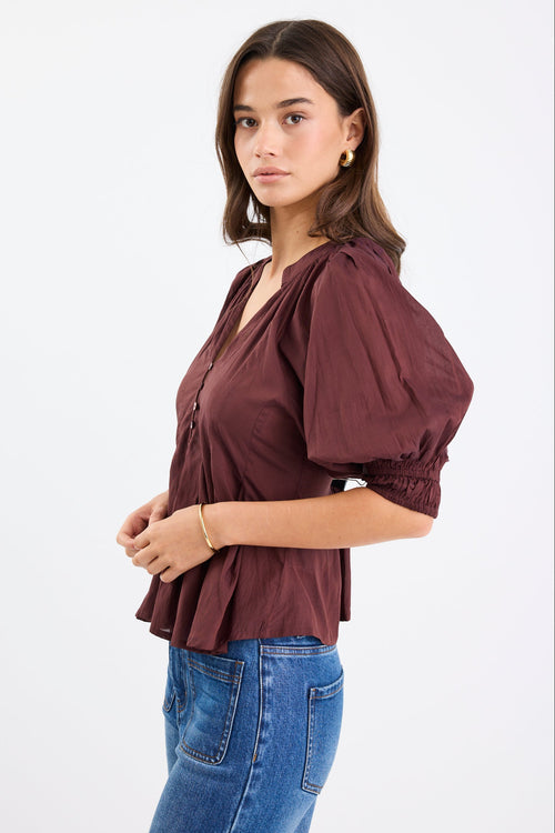 Woman wearing a chocolate brown pleated short-sleeve top with a button-down front, paired with blue jeans.