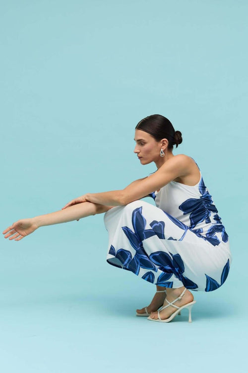 Woman wearing a sleeveless white midi dress with large blue floral print, featuring a side cutout detail and slim fit through the waist.
