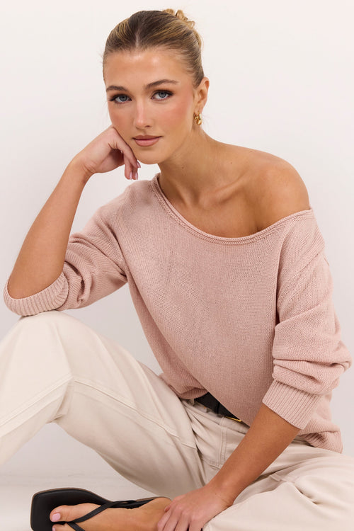 A woman wearing a dusty pink off-the-shoulder knit sweater, cream-colored pants, and black flat sandals, posing against a clean white studio background.