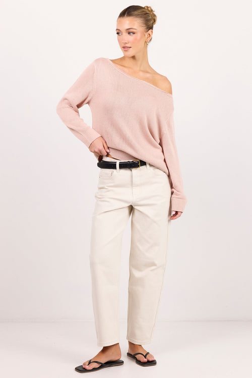 A woman modeling a light pink off-the-shoulder knit sweater, cream-colored straight-leg trousers, a black belt, and black sandals, standing against a seamless white studio background.