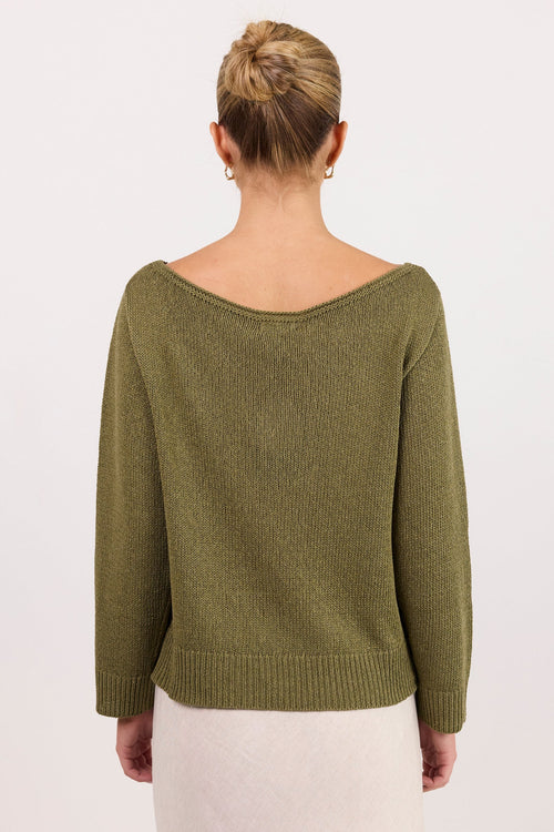 A woman viewed from the back wearing an olive green knit sweater with a wide boat neckline and a cream-colored skirt, set against a plain white studio background.