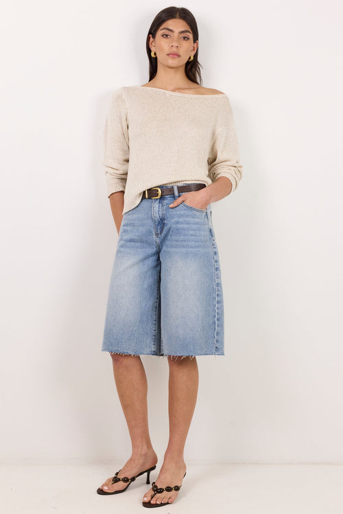 A woman modeling a light beige off-the-shoulder knit sweater and light-wash blue denim culottes with a dark belt, standing against a plain white studio background.