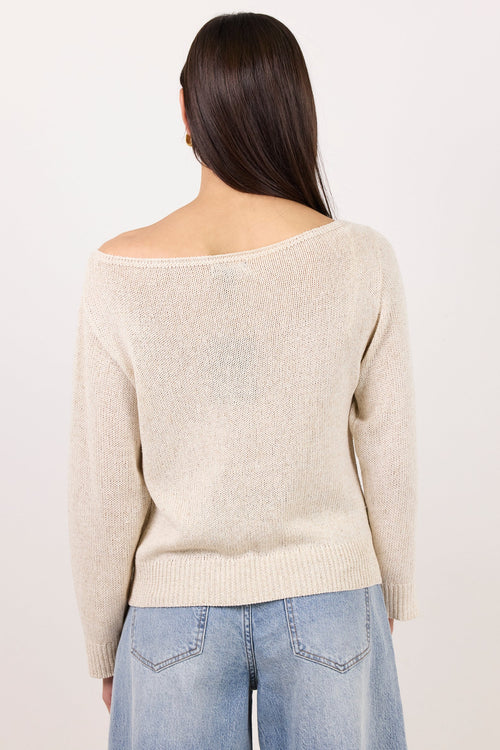 A woman viewed from the back wearing a light beige off-the-shoulder knit sweater and light blue jeans against a plain white studio background.