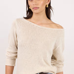 A woman wearing a cream-colored, off-the-shoulder knit sweater with three-quarter sleeves, paired with light blue jeans and gold hoop earrings, against a plain white studio background.