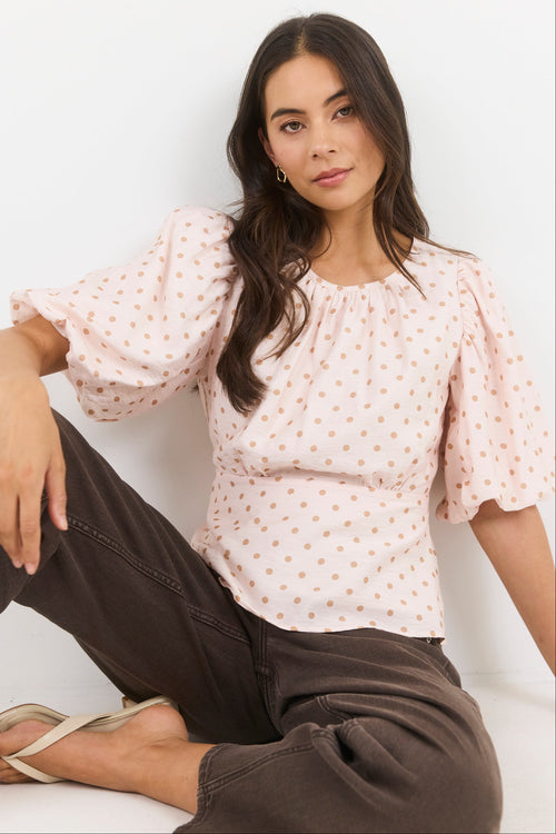 Woman wearing a light pink blouse with a subtle pattern and dark pants sitting on a white background