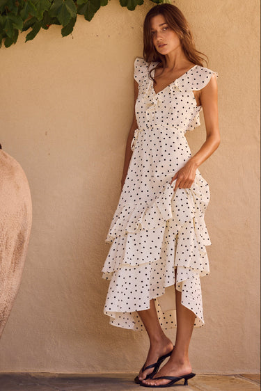 Woman in a white polka dot dress standing next to a large pot against a beige wall.