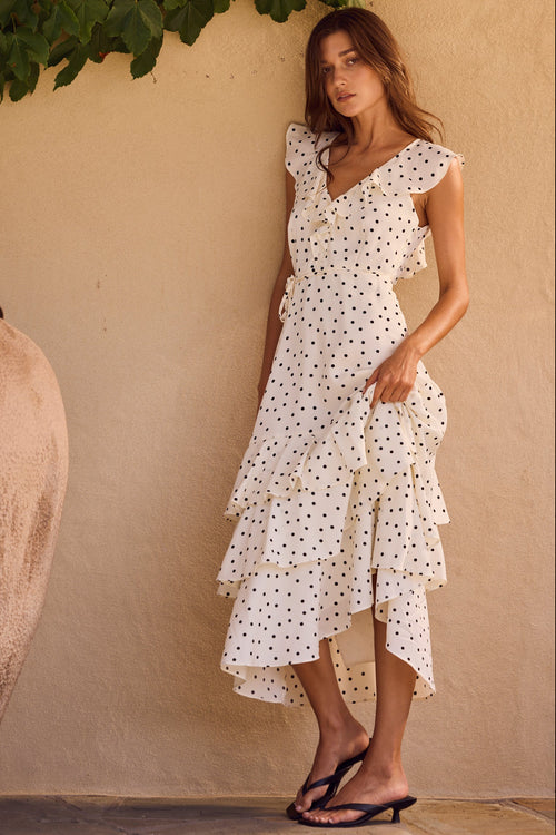 Woman in a white polka dot dress standing next to a large pot against a beige wall.