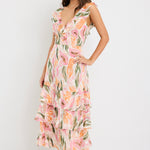 Woman wearing a sleeveless V-neck midi dress in a soft pink, peach, and green abstract print with a tiered skirt and tie detail at the back, styled with cream heeled sandals.
