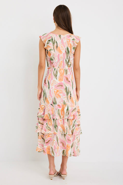 Woman wearing a sleeveless V-neck midi dress in a soft pink, peach, and green abstract print with a tiered skirt and tie detail at the back, styled with cream heeled sandals.