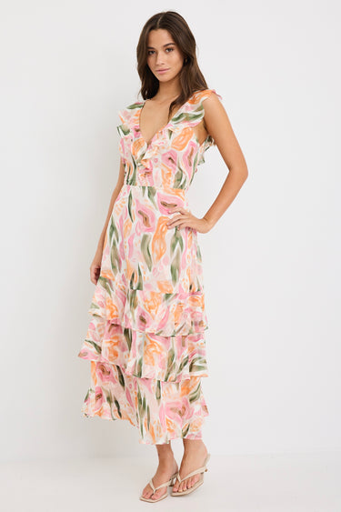 Woman wearing a sleeveless V-neck midi dress in a soft pink, peach, and green abstract print with a tiered skirt and tie detail at the back, styled with cream heeled sandals.