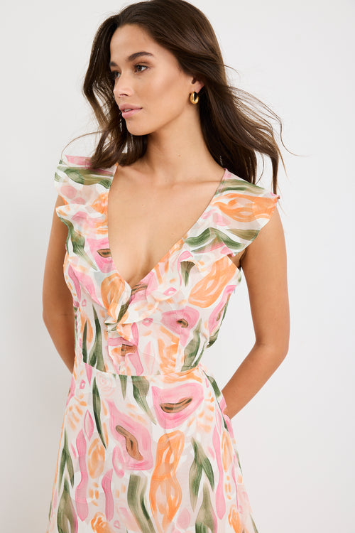 Woman wearing a sleeveless V-neck midi dress in a soft pink, peach, and green abstract print with a tiered skirt and tie detail at the back, styled with cream heeled sandals.