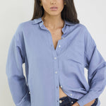 Woman wearing a light blue button-up shirt and dark jeans against a plain background