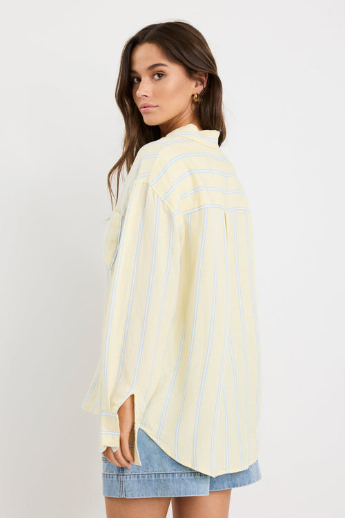Woman wearing an oversized yellow and blue striped button-front shirt with chest pocket, styled with mid-blue denim shorts.