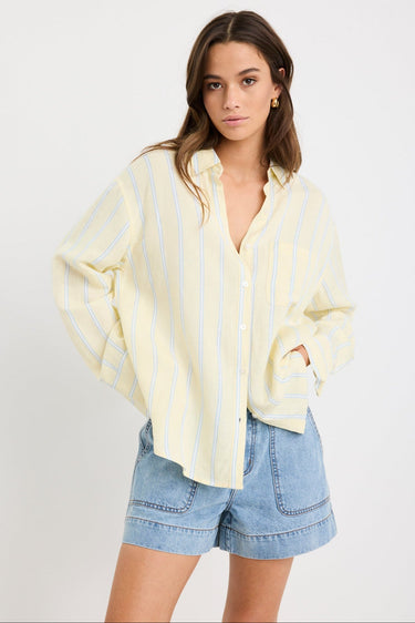 Woman wearing an oversized yellow and blue striped button-front shirt with chest pocket, styled with mid-blue denim shorts.