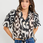 Woman wearing a black and cream zebra print short sleeve shirt with puff sleeves, button front, and tie waist detail, styled with blue jeans.