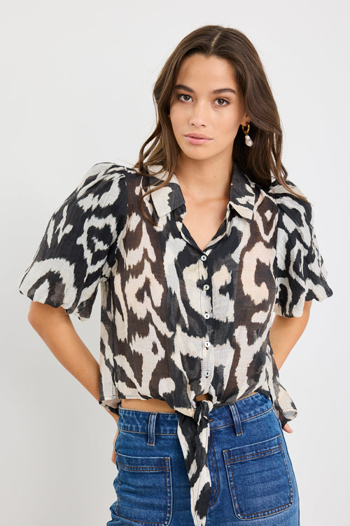 Woman wearing a black and cream zebra print short sleeve shirt with puff sleeves, button front, and tie waist detail, styled with blue jeans.
