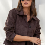 Woman wearing a brown jacket and blue jeans against a white wall.