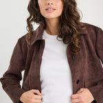 Woman wearing a brown suede jacket over a white shirt and blue jeans on a plain background
