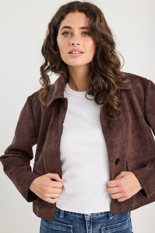 Woman wearing a brown suede jacket over a white shirt and blue jeans on a plain background